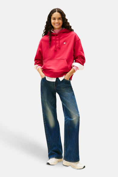 Pull-over rouge avec capuche de TOMMY JEANS, porté sur une veste rose et un chemisier blanc, avec des jeans bleus.