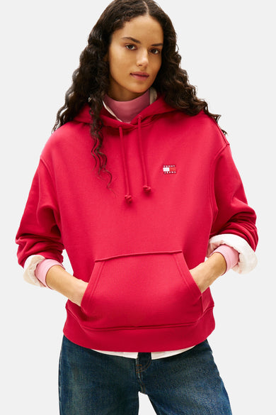 Pull-over rouge avec capuche de TOMMY JEANS, avec des cordons et une poche à l'avant, porté sur un gilet rose et un pantalon en cuir.