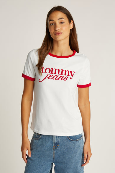 T-shirt blanc à manches courtes de TOMMY JEANS, avec une patte rouge et du texte coordonné. Porté avec des jeans bleus.