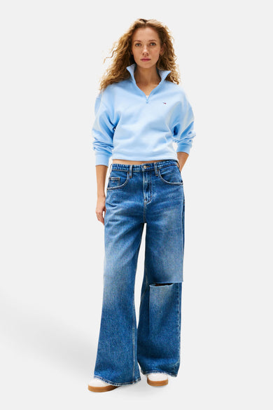 Pull-over bleu de TOMMY JEANS, avec une fermeture éclair courte et porté avec des jeans larges déchirés.