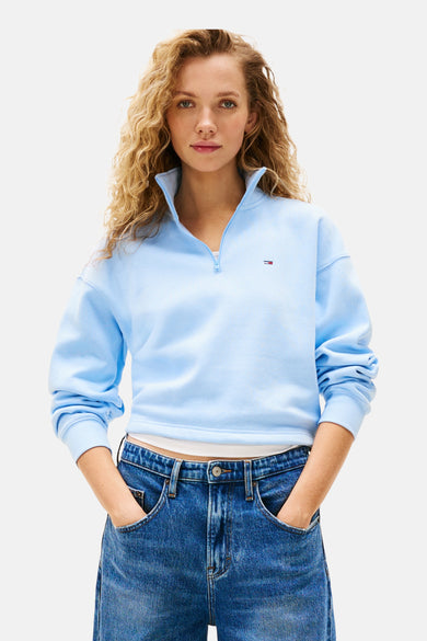 Pull-over bleu de TOMMY JEANS, avec une fermeture à glissière courte, porté avec un jeans bleu à larges jambes.