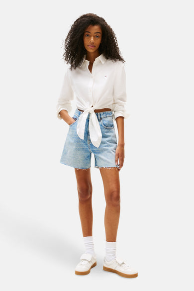 Chemise blanche à manches longues de TOMMY JEANS, boutonnée à l'avant, portée avec des shorts en denim et des baskets blanches.