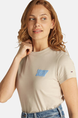 T-shirt écru à manches courtes de Tommy Jeans, avec une impression bleue sur la poitrine, porté avec un jean.