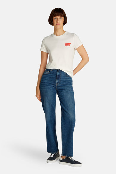 T-shirt blanc à manches courtes de TOMMY JEANS, avec du texte rouge sur la poitrine, associé à des jeans bleus et des baskets noires.