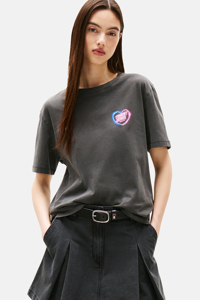 T-shirt gris à manches courtes de TOMMY JEANS, avec un logo en forme de cœur néon sur la poitrine et porté avec un pantalon noir et une ceinture.