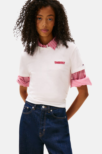 T-shirt blanc à manches courtes de TOMMY JEANS, porté sur un chemisier rayé avec un jean bleu foncé.