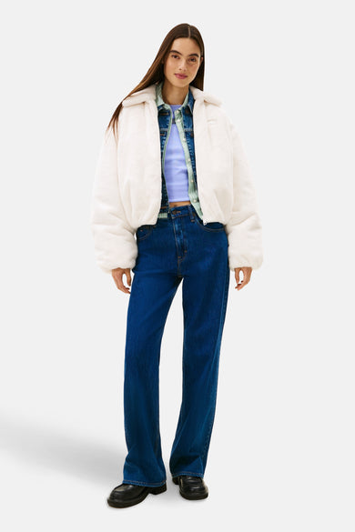 Manteau court blanc de TOMMY JEANS, porté sur une veste en cuir et un t-shirt violet, avec des jeans foncés et des chaussures noires.
