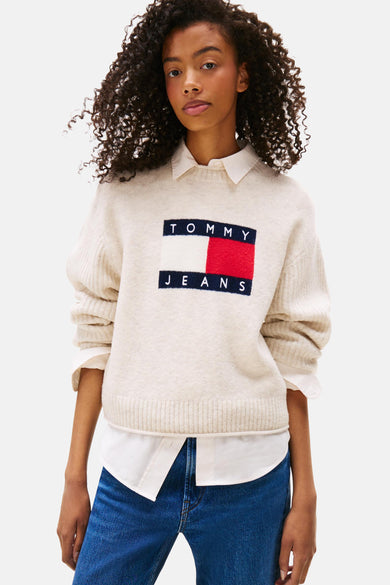 T-shirt écru avec col rond de TOMMY JEANS, avec un grand logo, porté sur une chemise blanche et des jeans bleus.