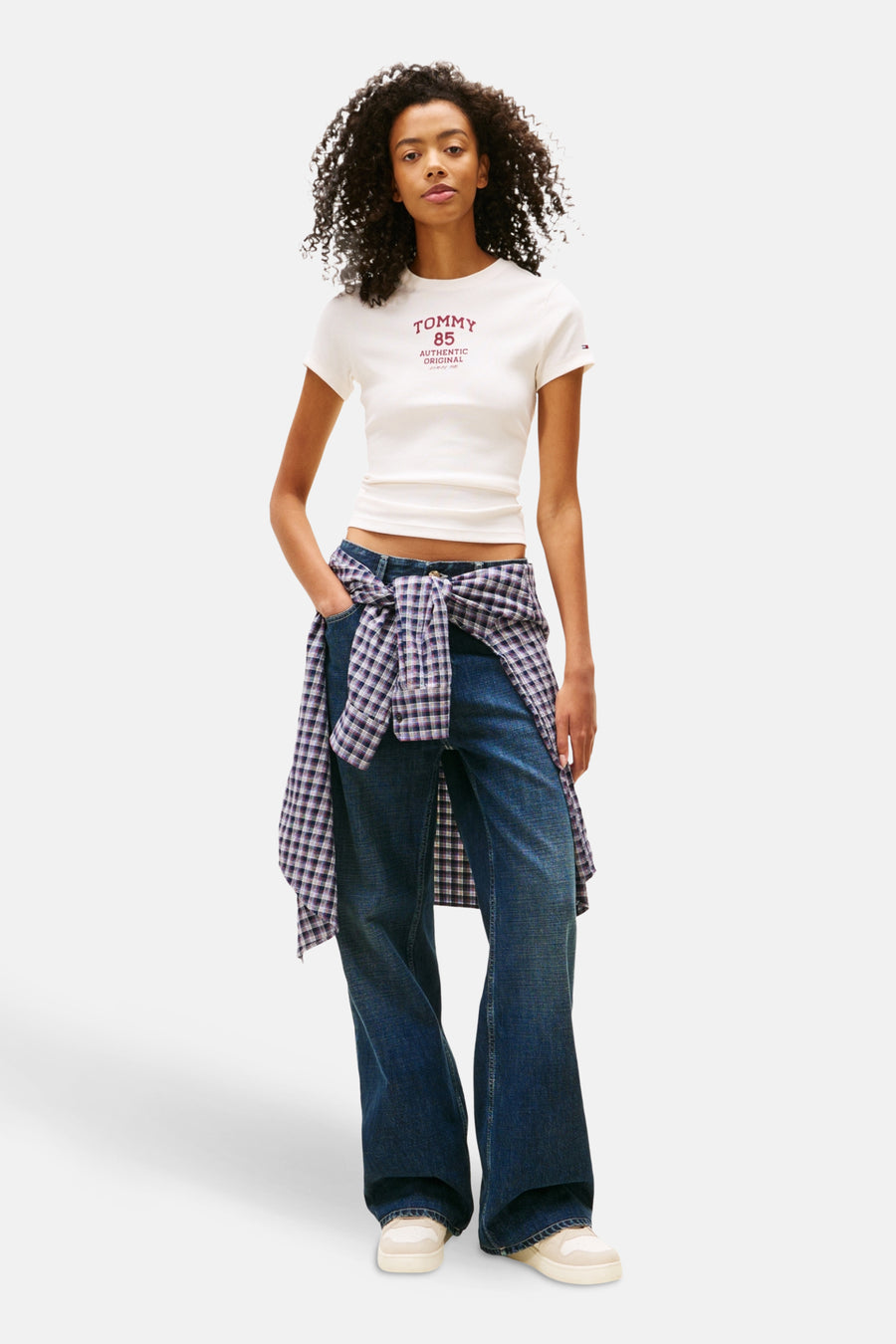 Wit T-shirt met korte mouwen van TOMMY JEANS, met rode tekst, gecombineerd met blauwe jeans en een geruit overhemd om de taille.
