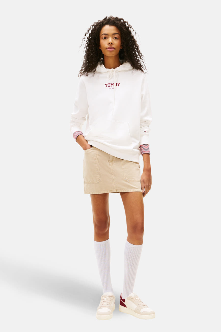 Witte sweater met kap van TOMMY JEANS, met gestreepte manchetten en gecombineerd met een beige rok, witte sokken en crèmekleurige sneakers.