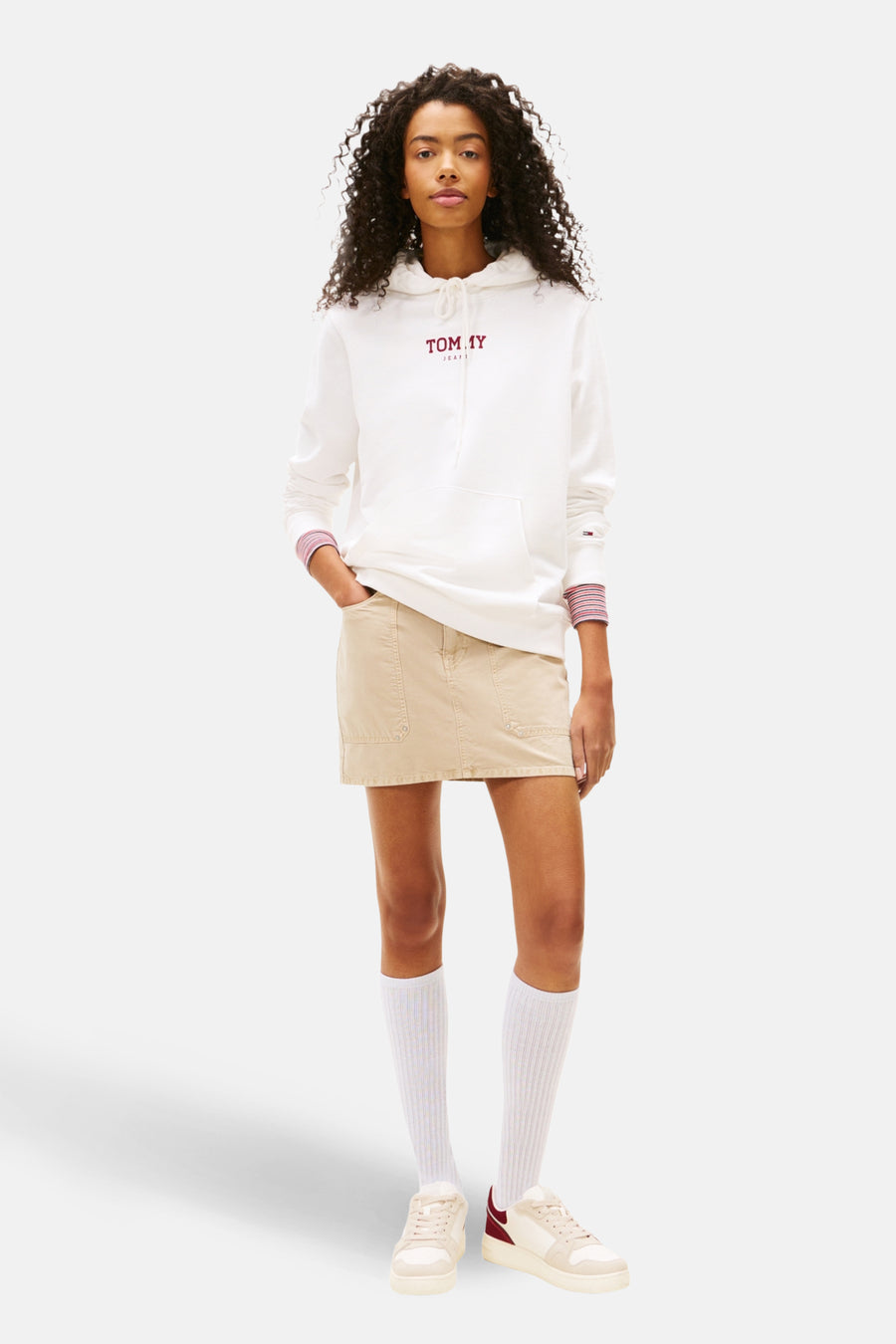Vrouw met krullend haar, witte Tommy Jeans hoodie, beige rok, witte sokken, en crèmekleurige sneakers.