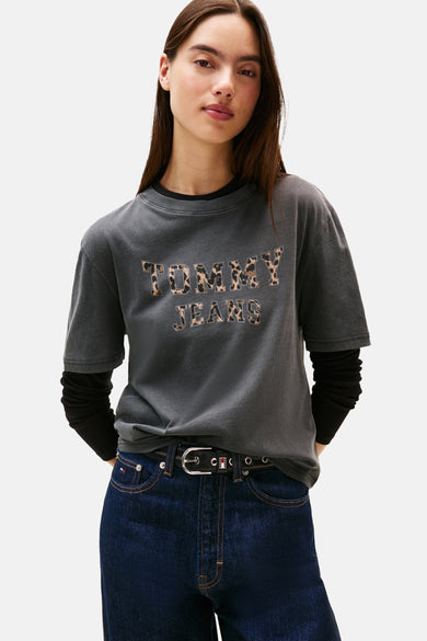 T-shirt gris à manches courtes de TOMMY JEANS, avec des lettres imprimées en léopard, associé à des jeans foncés et une ceinture noire.
