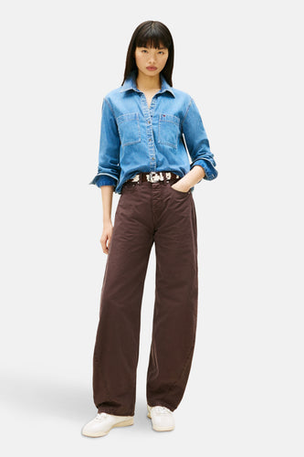 Vrouw in denim blouse en bruine wijde broek met riem, staand.
