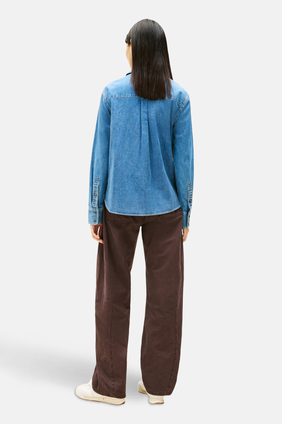 Achterkant van een vrouw in een spijkerblouse en bruine broek, op een witte achtergrond.