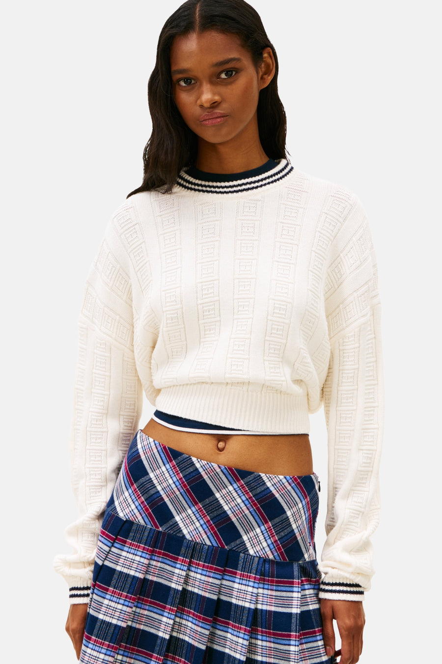 Model in een witte trui en een blauw-rood geruite plooirok.