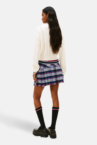 Achteraanzicht van een vrouw in een witte trui, geruite rok, zwarte sokken en leren schoenen.