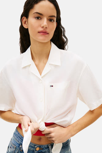 Chemise blanche à manches courtes TOMMY JEANS, nouée sur le devant, avec poche poitrine et portée avec un haut rouge et un jean.
