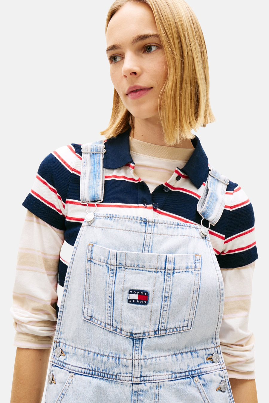 Combinaison en denim bleu clair de TOMMY JEANS, portée sur un polo rayé et un t-shirt à manches longues rayé, avec écusson logo sur la poitrine.