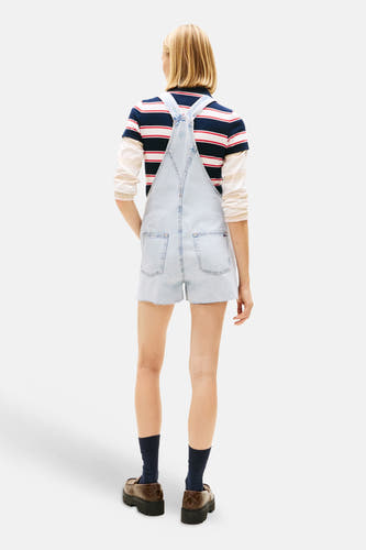 Combinaison en denim bleu clair de TOMMY JEANS, dos visible, portée sur une chemise rayée, avec des chaussettes bleu marine et des mocassins marron.
