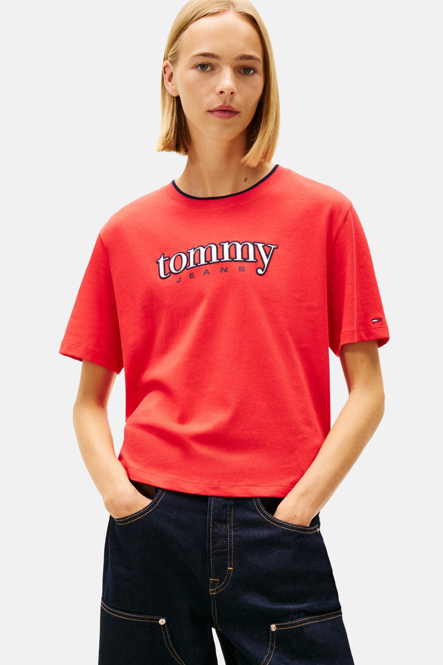 Model met blonde bob in een rode Tommy Jeans T-shirt en donkerblauwe broek, handen in de zakken, op een witte achtergrond.