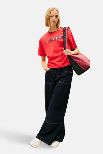 Een vrouw in een rode Tommy Jeans T-shirt, wijde donkerblauwe jeans en witte sneakers, met een geruite tas over haar schouder, poserend tegen een witte achtergrond.