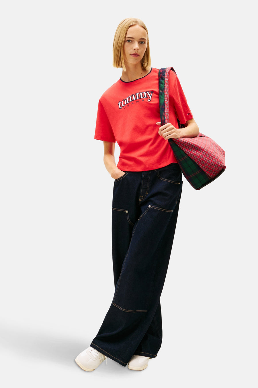 Een vrouw in een rode Tommy Jeans T-shirt, wijde donkerblauwe jeans en witte sneakers, met een geruite tas over haar schouder, poserend tegen een witte achtergrond.
