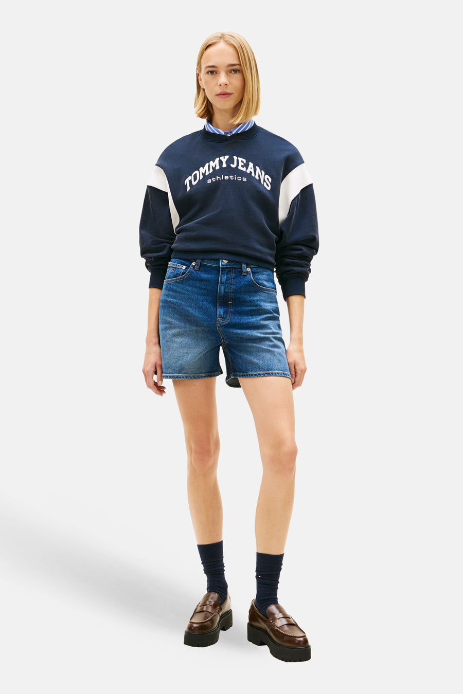 Donkerblauwe denim short van TOMMY JEANS, gedragen met een donkerblauw sweatshirt met witte details en bruine loafers met sokken.