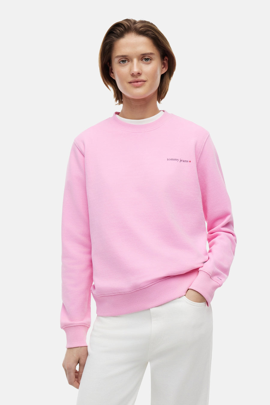 Pull rose à col rond de TOMMY JEANS avec logo rouge 'tommy jeans' sur la poitrine et drapeau sur la manche, porté par un mannequin.