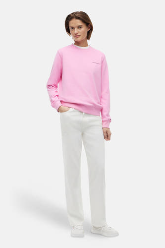 Pull rose à col rond de TOMMY JEANS, avec le texte 'tommy jeans' sur la poitrine, logo drapeau sur le poignet, et stylisé avec un jean blanc et des baskets.