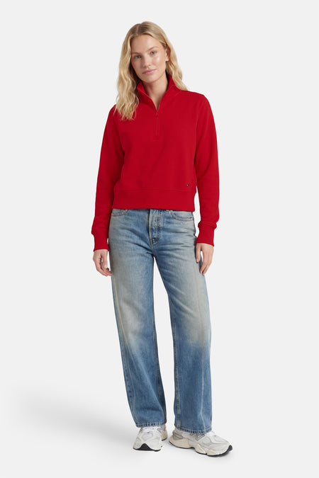 Rode ronde hals sweater van TOMMY JEANS, met halve rits en ribboorden, gedragen met lichtblauwe jeans en witte sneakers, front view.