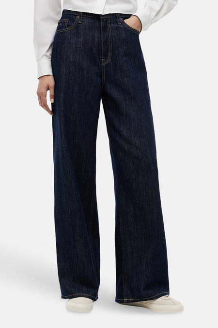 Donkere blauwe wijde jeans van TOMMY JEANS, gemaakt van denim, met hoge taille en rechte pijpen, gedragen met wit overhemd en crèmekleurige sneakers, front view.