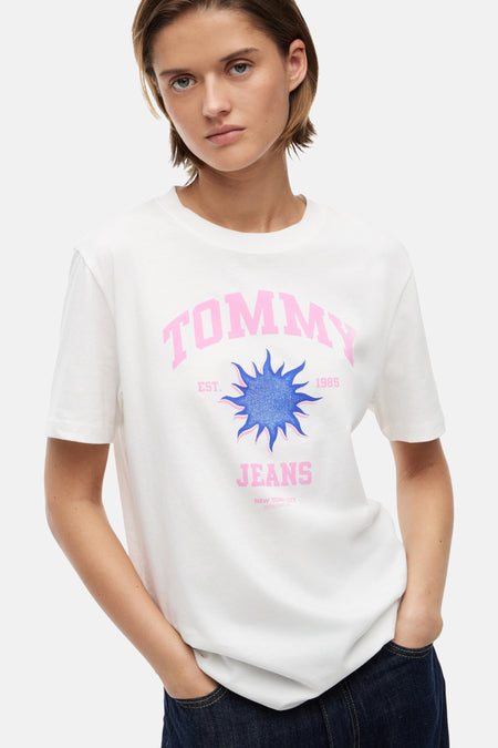Ecru top met korte mouwen van TOMMY JEANS met roze tekst, een blauwe zongrafiek en een model in donkere jeans.