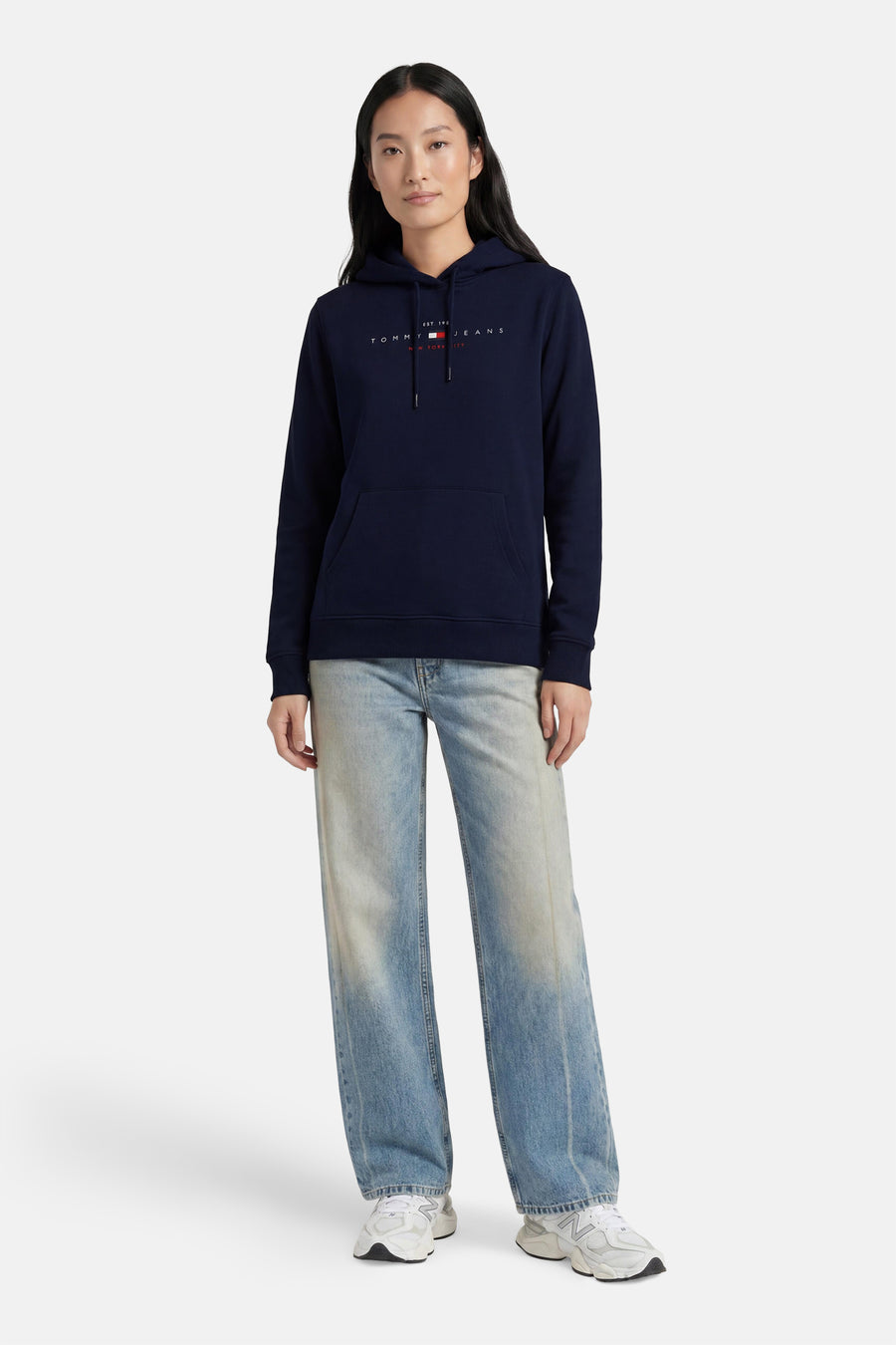 Donkerblauwe sweater met kap van TOMMY JEANS, voorzien van logo op borst, met zak en trekkoord, gedragen met lichte jeans en witte sneakers.