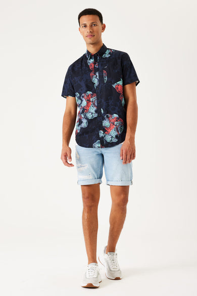 Chemise bleue à manches courtes de GARCIA, avec un motif floral frappant en rouge et bleu, associée à des shorts en denim et des baskets blanches.
