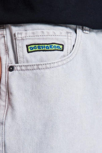 Lichtgrijze denim jeans met speciale pasvorm van DC SHOES, close-up van de voorzak met geborduurd logo en zwarte rivet.