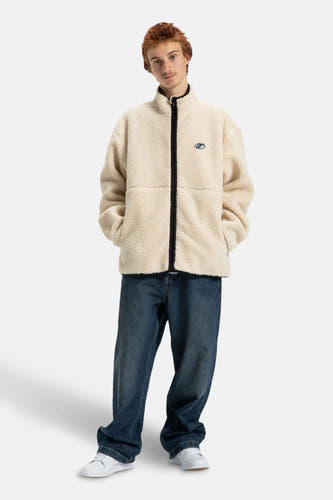 Manteau court beige de DC SHOES, avec des manches longues et porté avec un jeans bleu et des chaussures blanches.