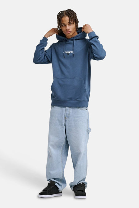 Sweat à capuche bleu de DC Shoes, avec cordons de serrage et une poche kangourou. L'homme porte un jean ample et des chaussures noires.