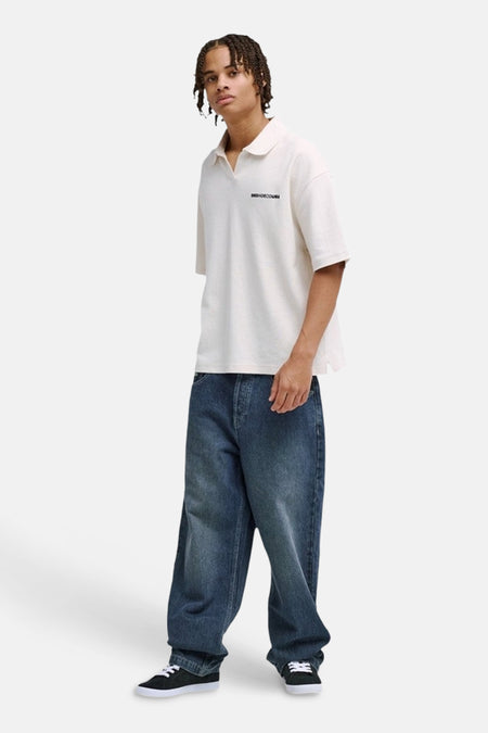 Polo blanche DC SHOES avec col ouvert et logo texte noir, portée par un homme avec un jean ample et des baskets noires.