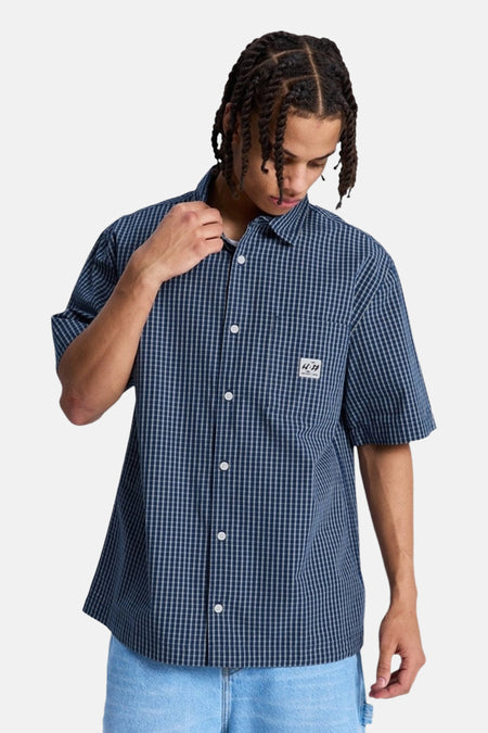 Chemise à carreaux bleus à manches courtes de DC SHOES, portée dans le dos par une personne avec des twists et un jean bleu clair.