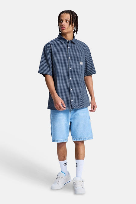 Chemise à carreaux bleus à manches courtes de DC SHOES, portée par un homme avec un short en jean et des baskets.