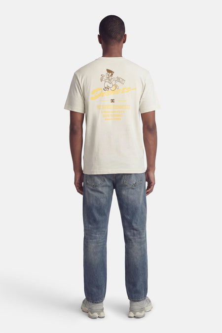 T-shirt beige de DC SHOES, avec un dessin animé et du texte au dos, porté avec un jean et des baskets grises.