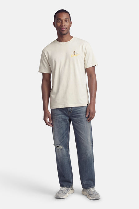 T-shirt beige de DC SHOES à manches courtes, coupe ample et petit imprimé sur la poitrine, porté avec un jean bleu.