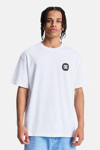 T-shirt blanc à manches courtes de DC SHOES, avec un logo noir et blanc sur la poitrine gauche, porté par un homme aux cheveux tressés et un jean bleu.