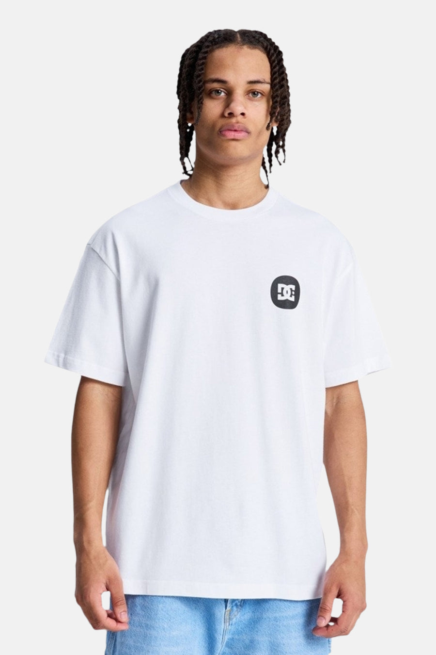 T-shirt blanc à manches courtes de DC SHOES, avec un logo noir et blanc sur la poitrine gauche, porté par un homme aux cheveux tressés et un jean bleu.