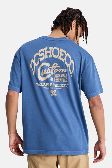 T-shirt bleu à manches courtes de DC SHOES, avec un logo sur la poitrine, porté avec un pantalon kaki et des chaussures noires.