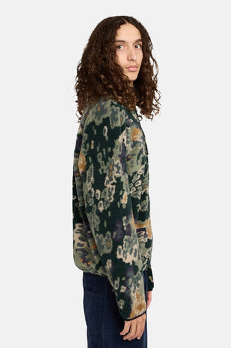 Un homme aux cheveux longs bouclés porte une chemise en fleece verte à motif floral et un pantalon bleu marine, debout en profil contre un fond blanc.
