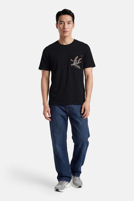 T-shirt noir ELEMENT avec une poche poitrine à imprimé abstrait dans des tons terreux, porté par un homme en jean bleu et baskets grises.