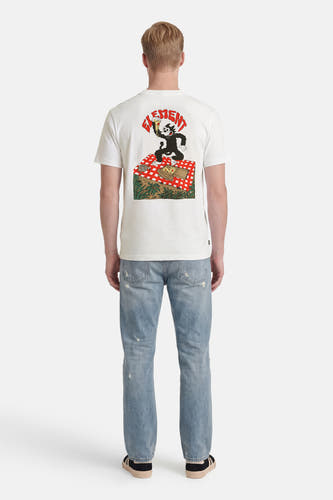 T-shirt blanc ELEMENT avec un imprimé de Felix the Cat sur une couverture de pique-nique, porté par un homme avec un jean délavé et des baskets, vu de dos.