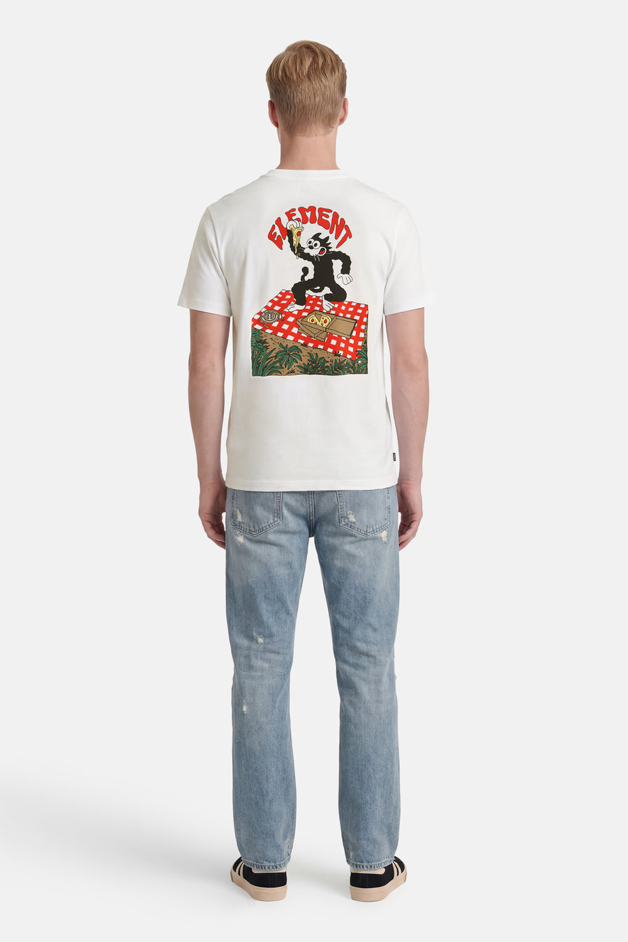 T-shirt blanc ELEMENT avec un imprimé de Felix the Cat sur une couverture de pique-nique, porté par un homme avec un jean délavé et des baskets, vu de dos.
