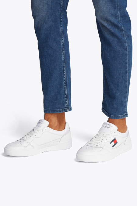 Sneakers blanches de TOMMY JEANS, avec logo en rouge, blanc et bleu, portées avec des jeans bleus.